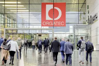 Menschen gehen zum Eingang eines großen Gebäudes mit einem roten Schild mit der Aufschrift "Orgatec".